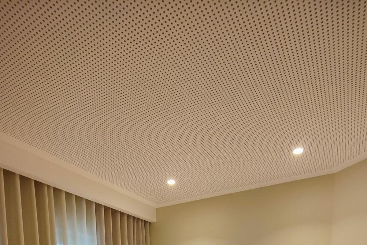 A hotel room with a wooden bed, a patterned bedspread, a headboard, a bedside table with a phone, and a ceiling with a perforated design.