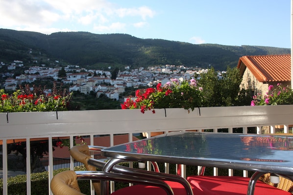 A balcony with a table and chairs, overlooking a town with red-roofed houses and a mountain in the background. - Hotel Berne (Manteigas)