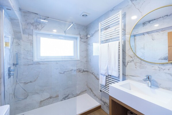 A modern bathroom with a glass shower enclosure, a built-in window, a white sink, and a marble-tiled wall. - Hotel Berne (Manteigas)