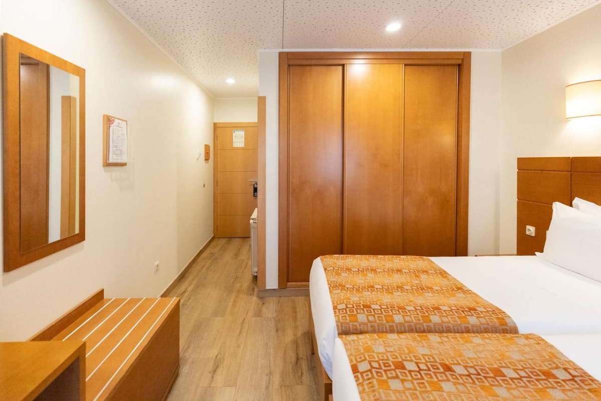 A hotel room with a wooden wardrobe, a bed with patterned bedding, a bedside table, and a mirror on the wall.