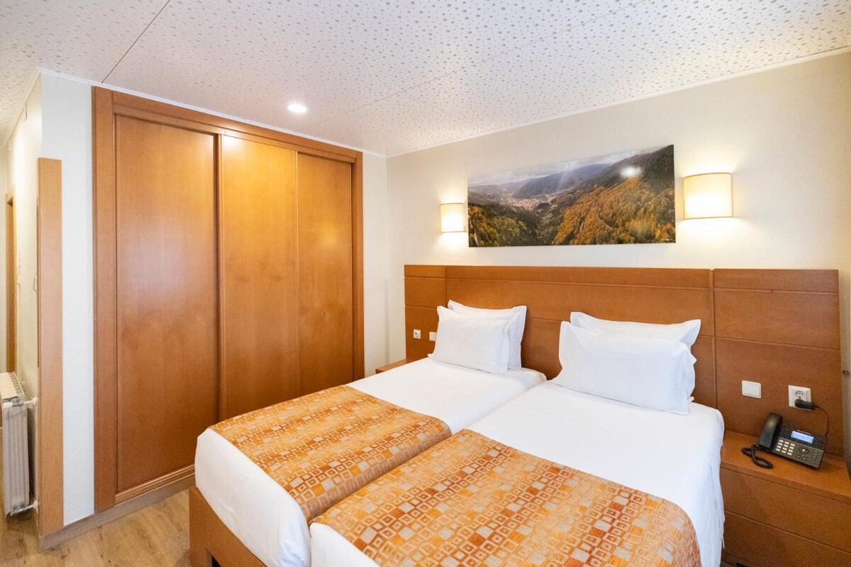 A hotel room with a double bed, a wooden wardrobe, a painting on the wall, and a bedside phone.