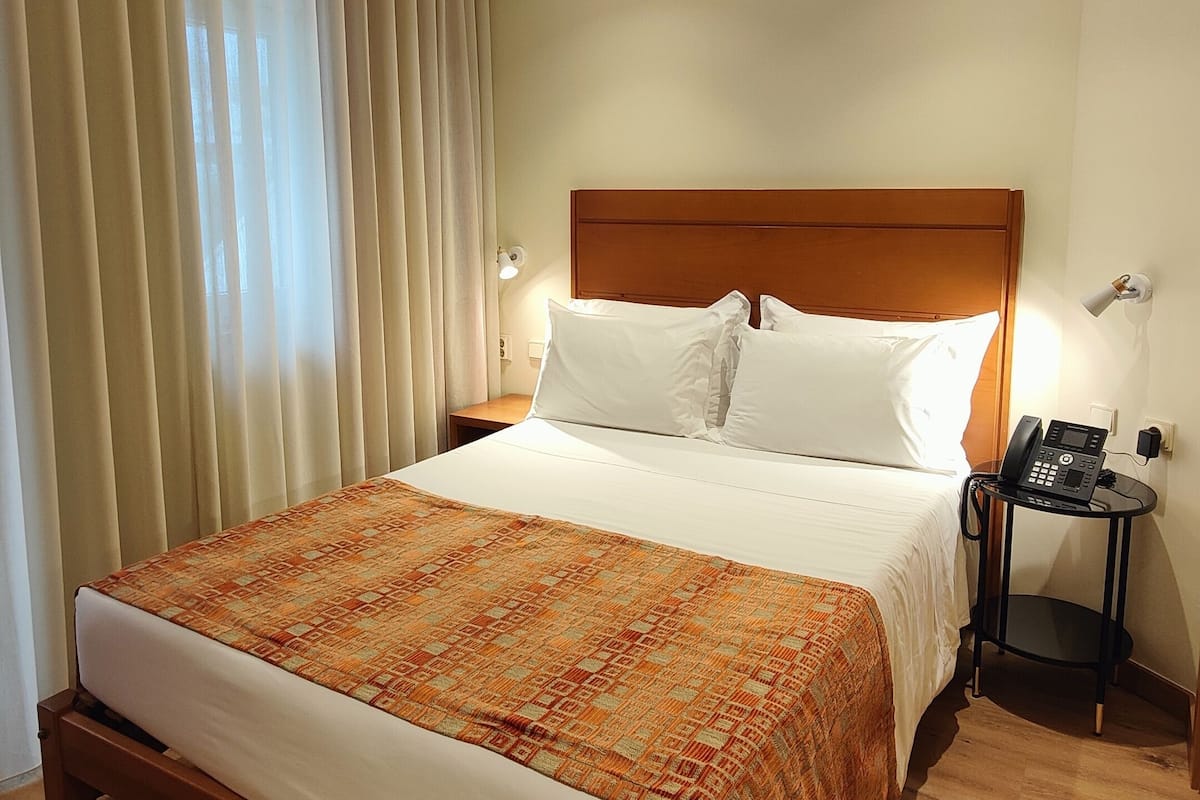 A hotel room with a wooden bed, a patterned bedspread, a wooden headboard, a bedside table with a phone, and a ceiling with a perforated design.
