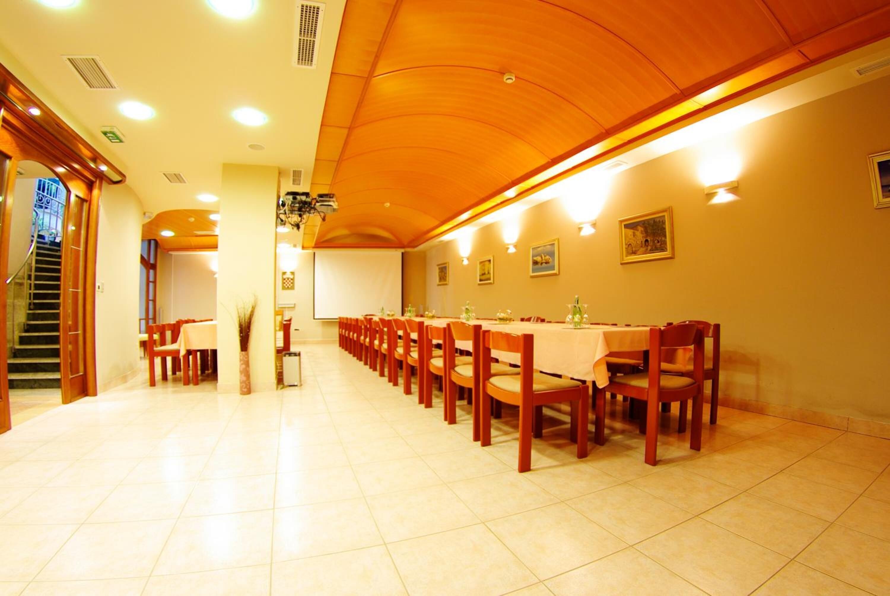 A spacious dining hall with wooden tables and chairs, a high ceiling, and a staircase leading to another area.