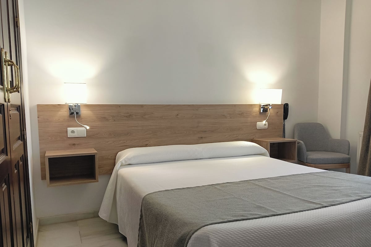 Una habitación de hotel con cabecera de madera, cama con ropa de cama blanca, un sillón gris y lámparas de noche.