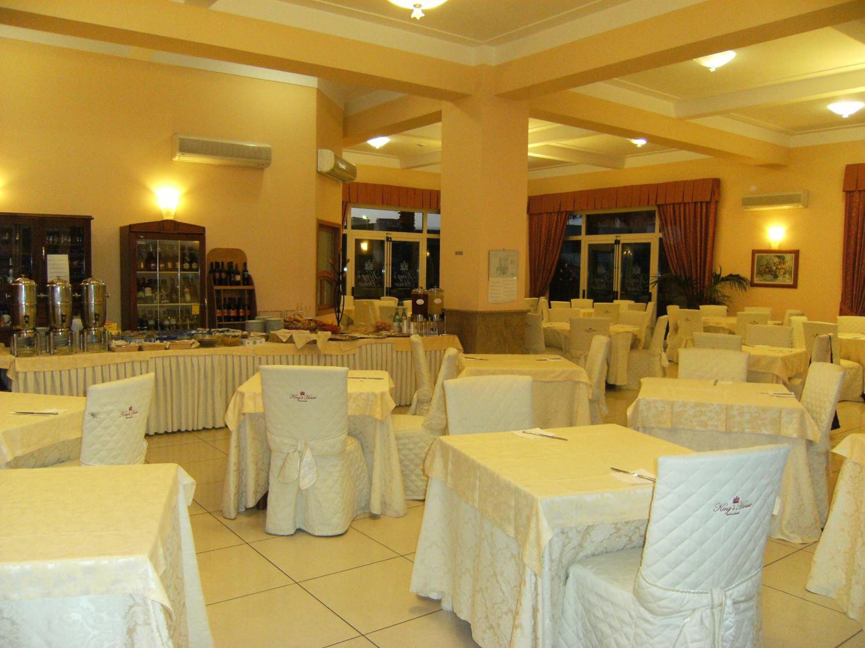 Une salle de banquet avec des tables rondes recouvertes de nappes blanches et des chaises, un espace buffet proposant des plats et des boissons, et une grande fenêtre ornée de rideaux.