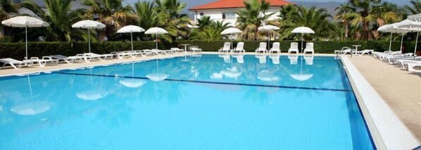 Una piscina con sillas de descanso blancas y sombrillas, rodeada de palmeras y un edificio con techo rojo.