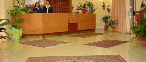 Un espace d’accueil avec un bureau en bois, deux réceptionnistes, un grand tapis à motifs et plusieurs plantes en pot.
