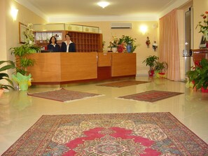A reception area with a wooden desk, two receptionists, a large patterned rug, and several potted plants. - King's House Hotel Resort (Mascali)