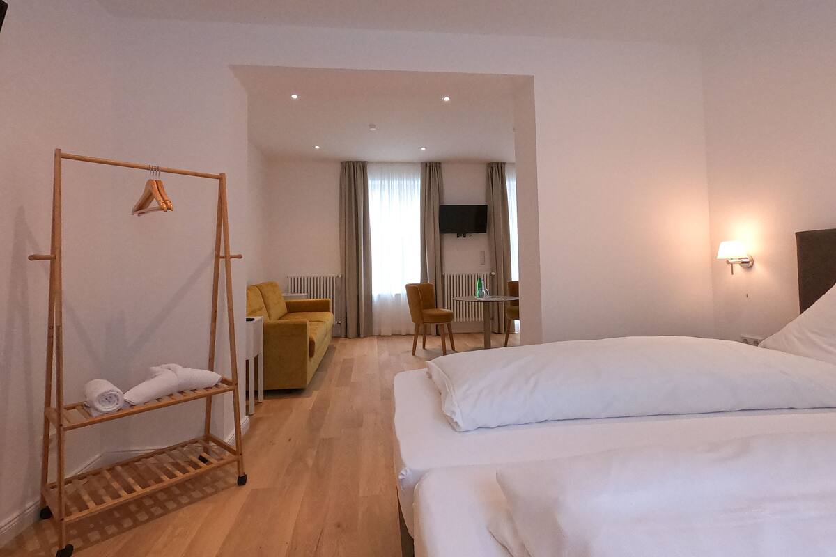 A hotel room with two beds, a wooden drying rack, a sofa, a dining table with chairs, a TV mounted on the wall, and a radiator.