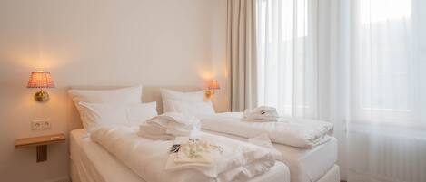 A hotel room with two beds, a nightstand, a lamp, and a window with sheer curtains.