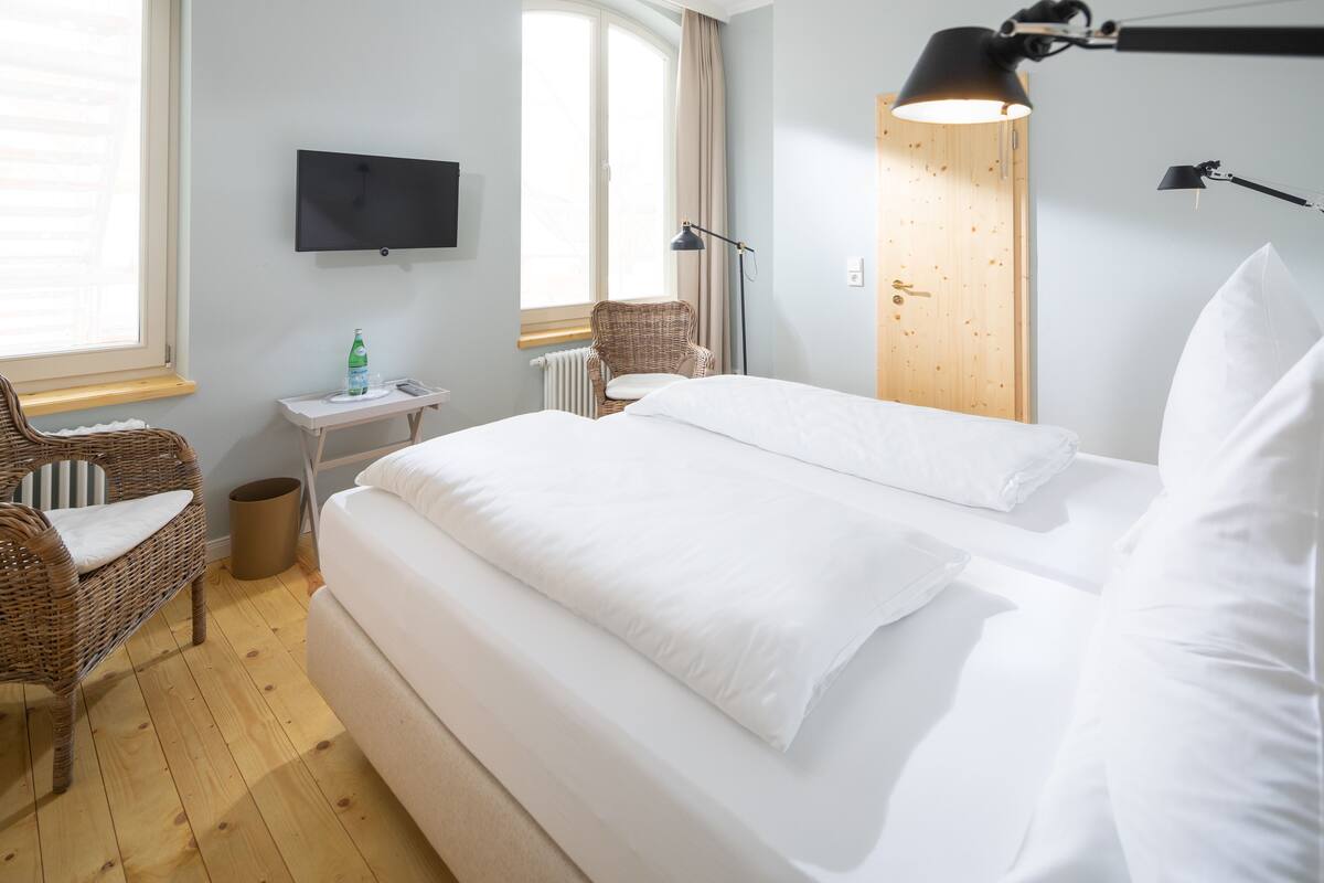 A hotel room with a bed, a chair, a small table, a TV, and a window with curtains.