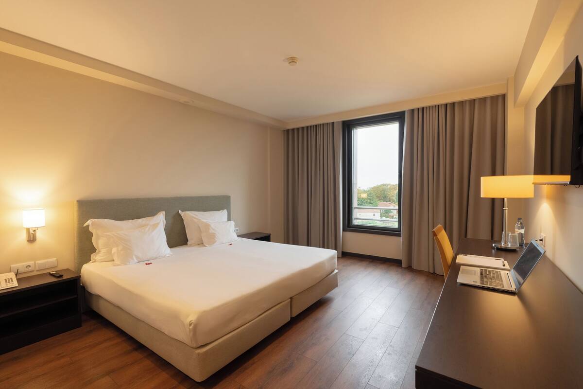 A hotel room with a large bed, a desk with a laptop, a telephone, and a window with curtains.