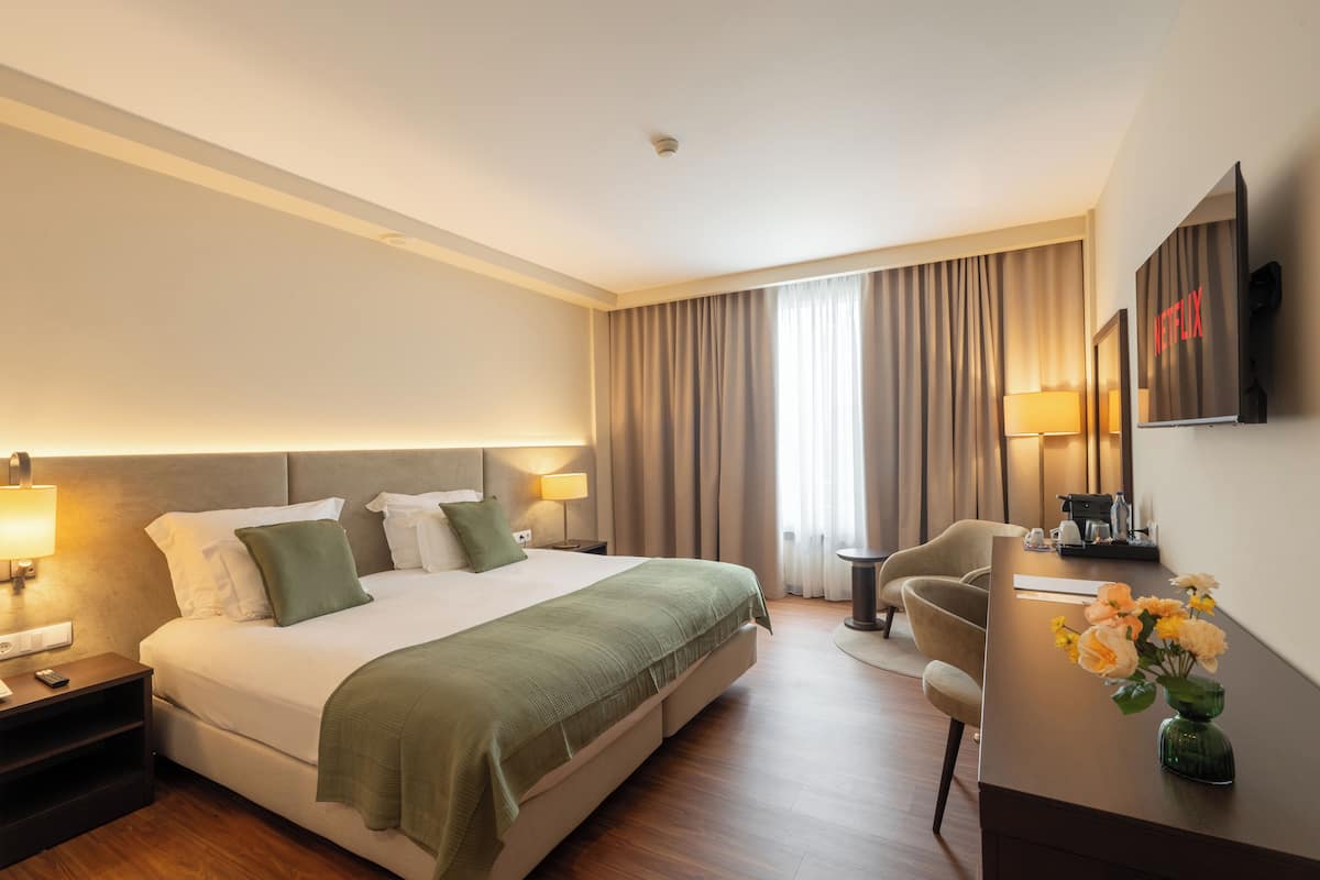 A hotel room with a bed, bedside lamps, a desk with a chair, a television, and a vase with flowers.
