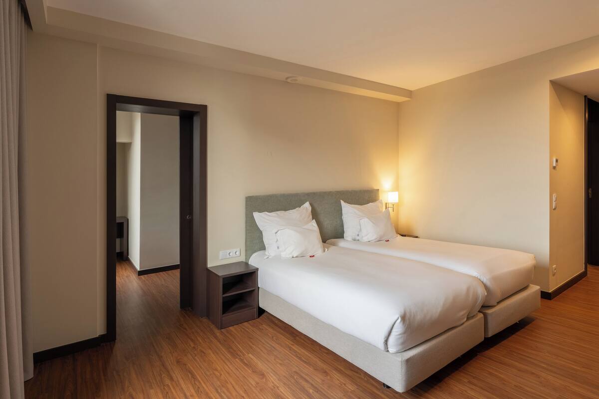 A hotel room with two beds, a nightstand, and a door leading to another room.