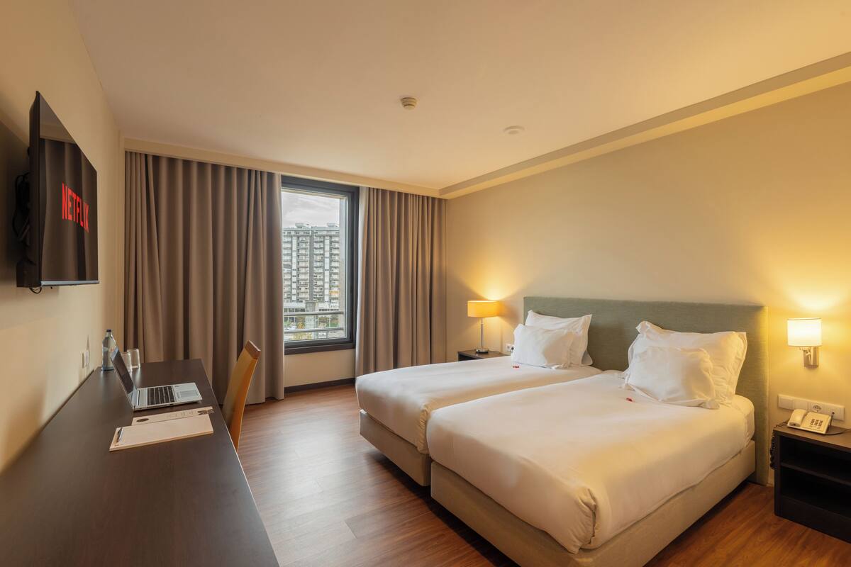 A hotel room with two beds, a desk with a laptop, a TV, and a window with curtains.