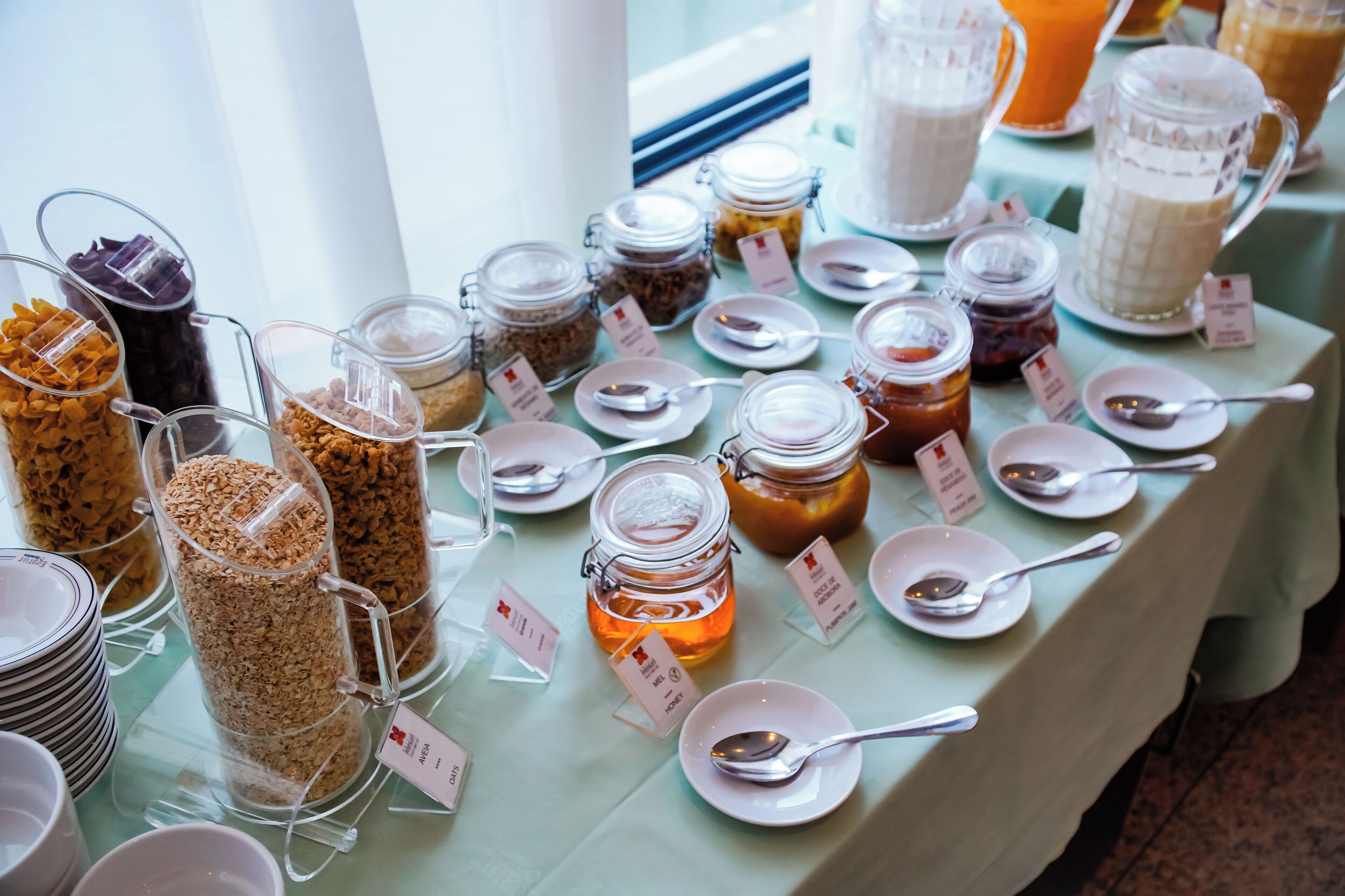 A buffet table with various food items and condiments, including jars of pickles, honey, and jams, with serving utensils and labels.