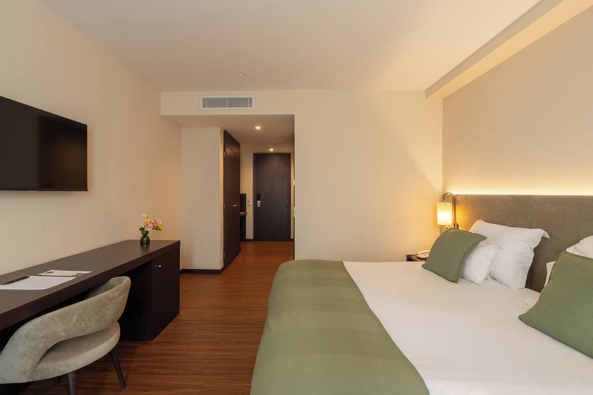 A hotel room with a bed, a desk, a chair, a flat-screen TV, and a nightstand with a lamp.