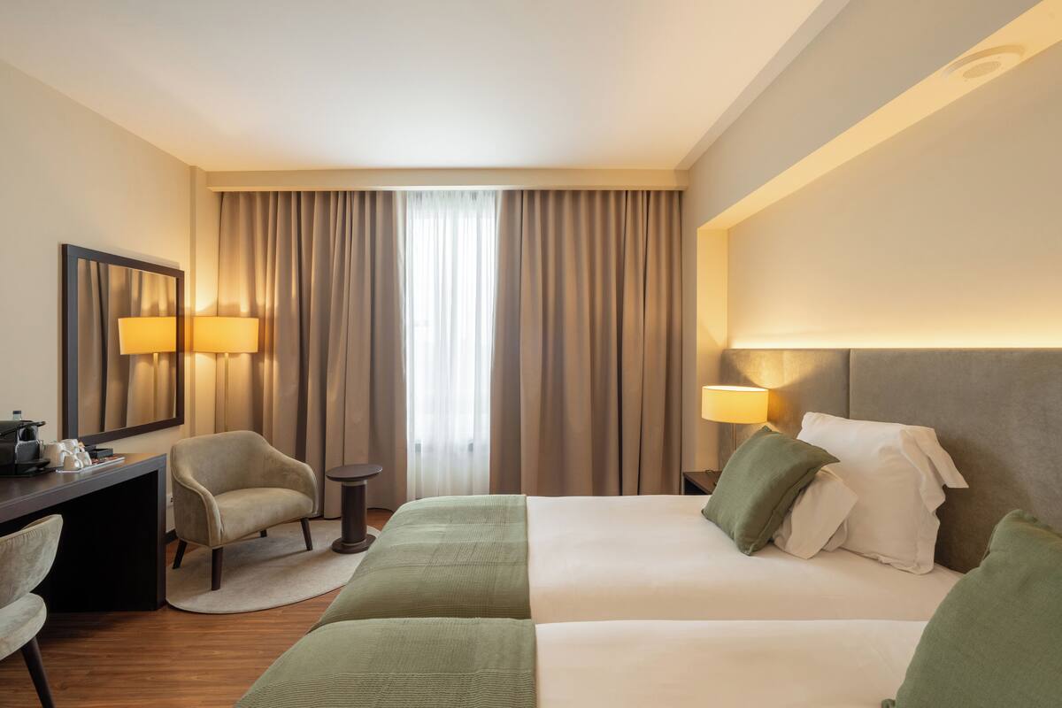 A hotel room with a bed, two chairs, a desk, a lamp, and a large window with curtains.