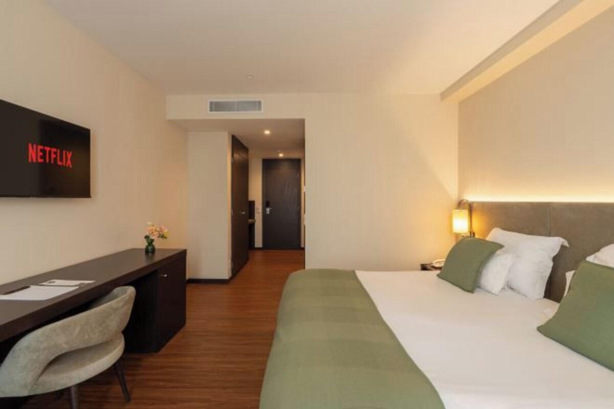 A hotel room with a bed, a desk, a chair, a TV screen displaying 'NETFLIX', and a door leading to another room.