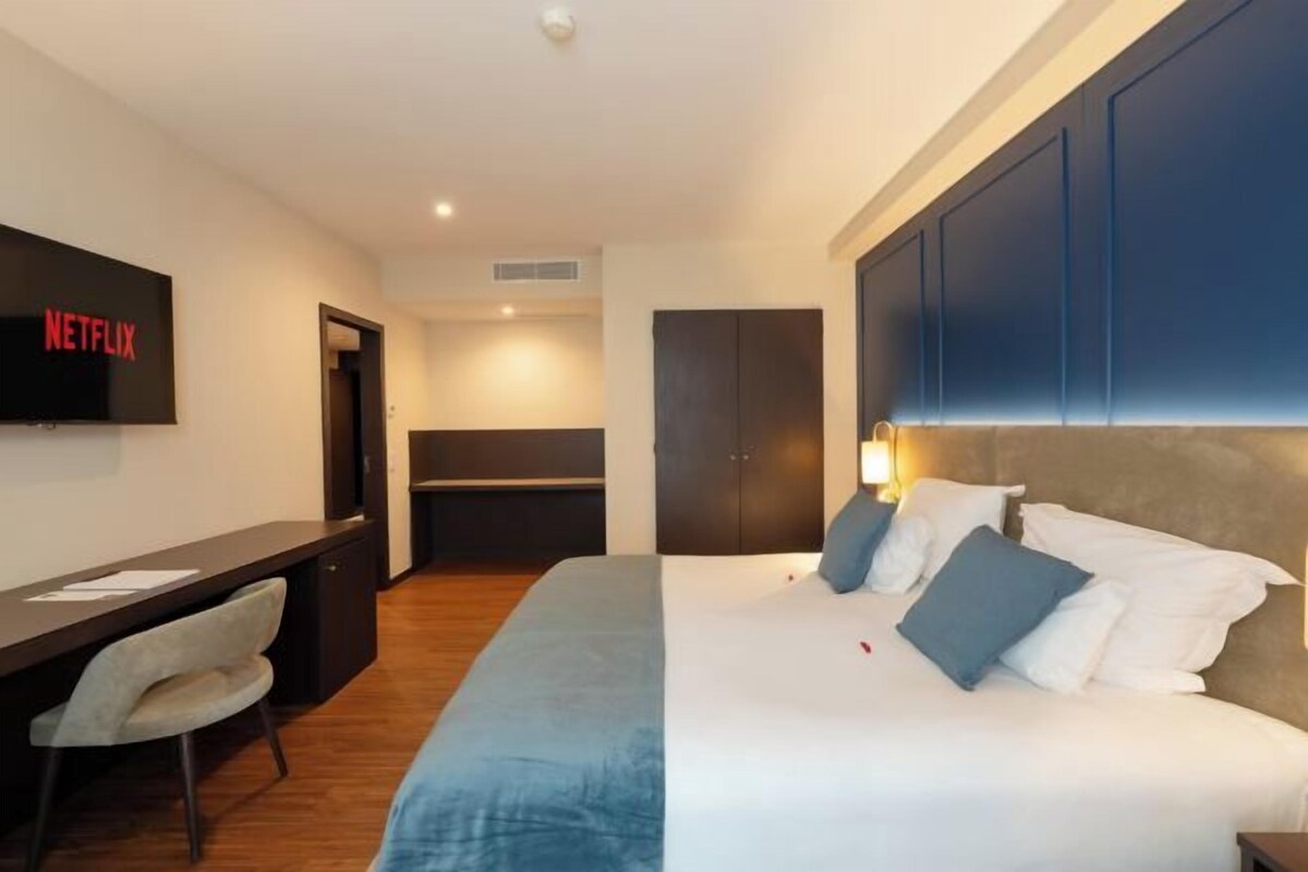 A hotel room with a bed, a desk, a chair, a wardrobe, and a television displaying the Netflix logo.