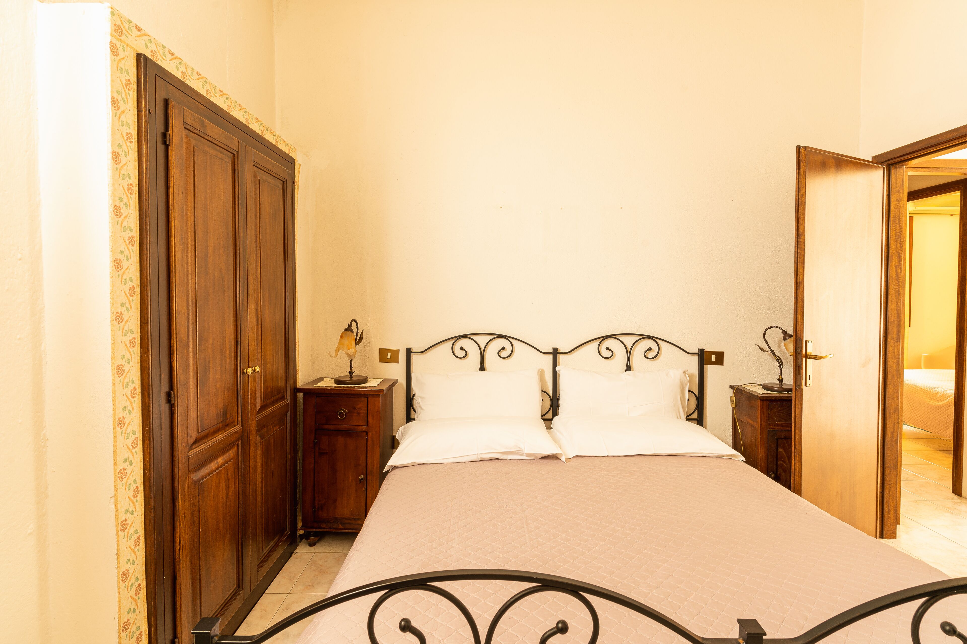 Un dormitorio con una cama, un armario de madera, dos mesitas de noche con lámparas y una puerta que da a otra habitación.