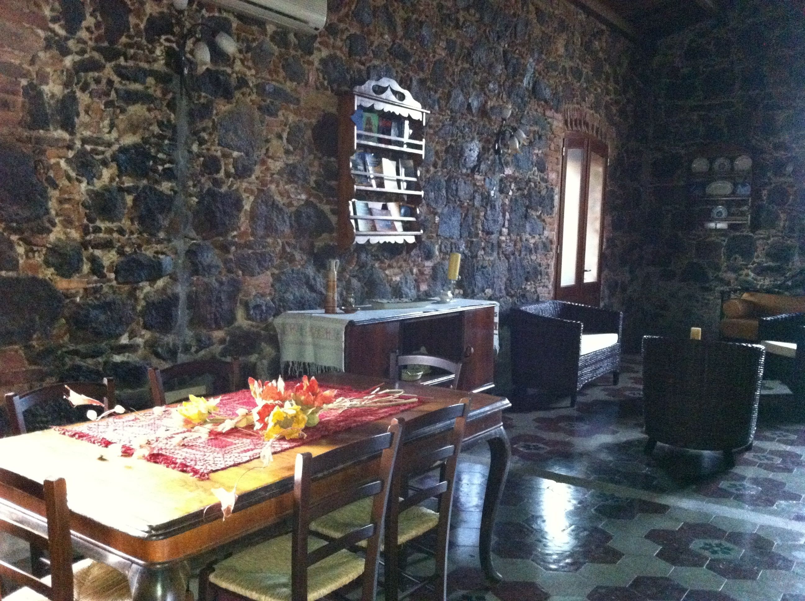 A stone-walled dining area with a table set for two, a wooden chair, and a small shelf with books.