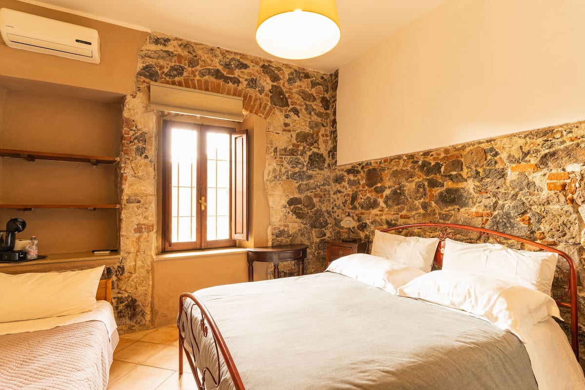 A bedroom with a stone wall, a bed with white linens, a bedside table, and a window with bars.