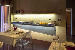 A breakfast bar with a variety of food items and beverages, a seating area with tables and chairs, and a wall-mounted shelf with kitchen utensils. - Hotel Remàt (Garda)