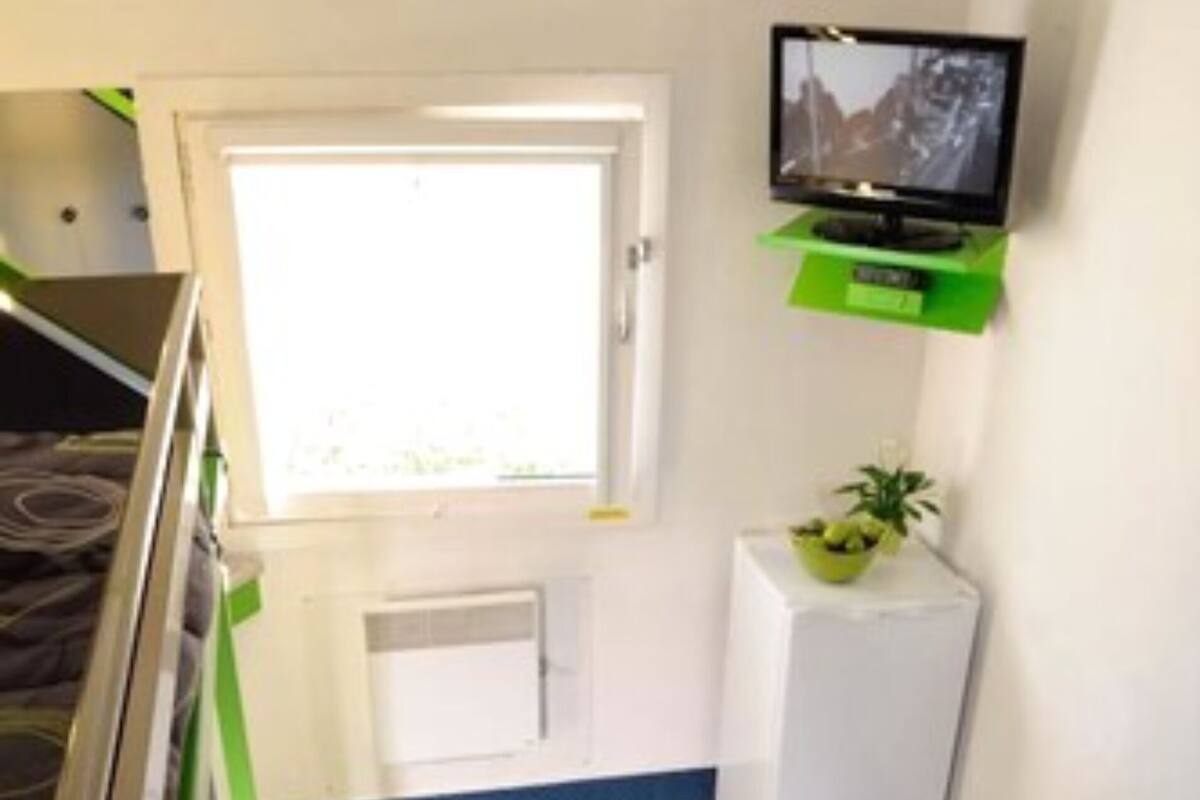 Een kamer met een bed, een televisie bevestigd aan een groen schap, een klein wit kastje en een raam.