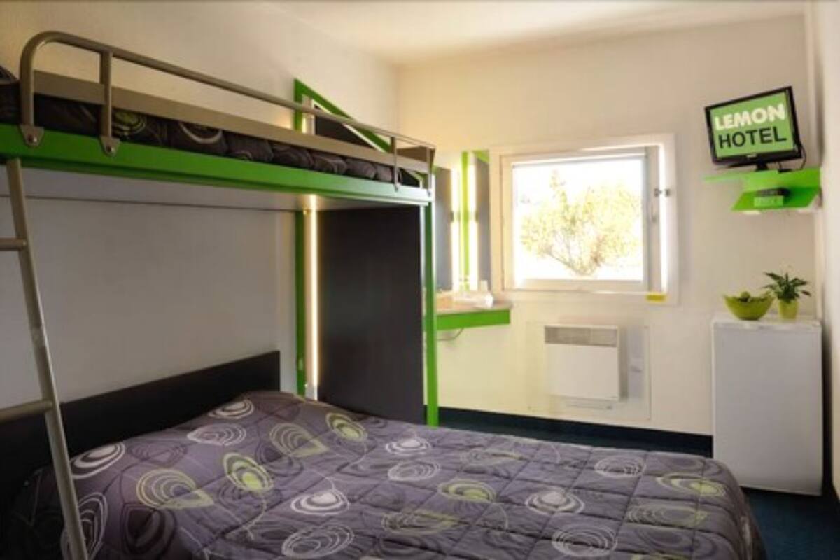 Een eenpersoonsbed met een bed boven, een klein bureau met stoel, een raam met uitzicht op bomen, en een bordje met 'LEMON HOTEL'.