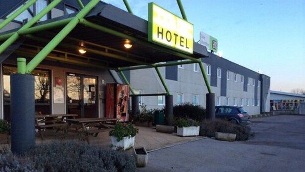 Un hôtel doté d’un espace extérieur couvert avec des sièges, d’une enseigne indiquant la présence d’un hôtel et d’un parking où se trouve une voiture.