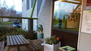 Une terrasse avec une table et des bancs en bois, un bac à fleurs blanc contenant une plante et une fleur rouge, et un chat assis par terre.