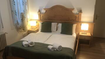 Ein ordentlich bezogenes Bett mit zwei Kissen, einer grünen Tagesdecke und zwei aufgerollten Handtüchern. Auf den Holznachttischen stehen jeweils zwei Nachttischlampen, und links ist ein Heizkörper zu sehen.