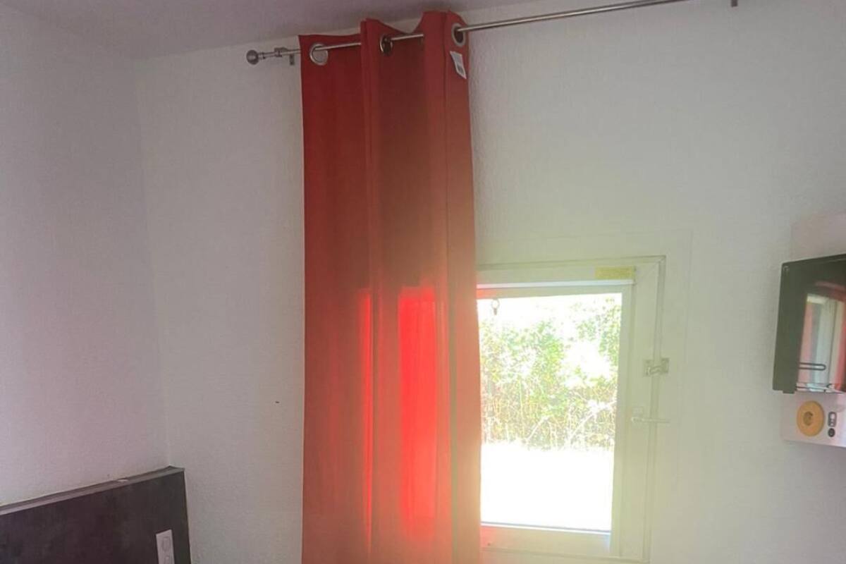 Une pièce avec un lit, une fenêtre avec un rideau et une étagère fixée au mur, où sont rangés des livres.