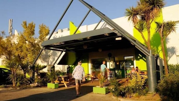 Un edificio moderno con estructura de techo triangular, zona de estar al aire libre y vegetación.