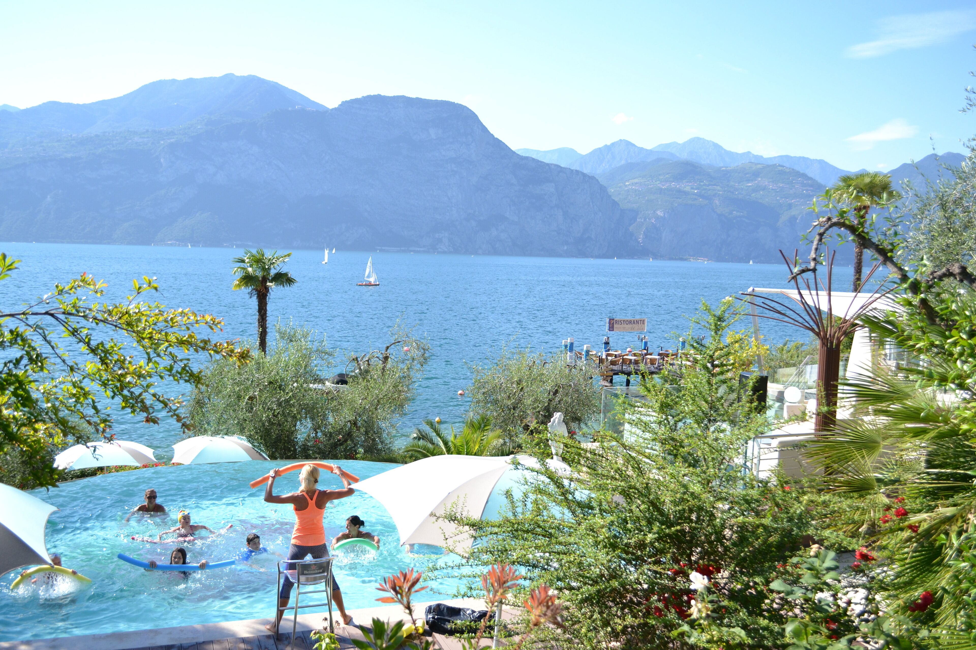 Ein Poolbereich mit Schwimmern und spielenden Personen, umgeben von üppiger Vegetation und mit Blick auf einen See mit Segelbooten.