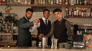 Een barsetting met drie bartenders, diverse drankflessen op planken en baraccessoires op het aanrecht.