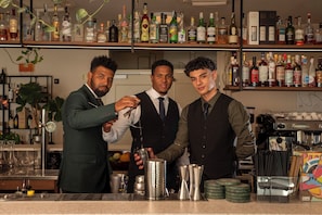 En bardisk med tre bartenders, ett urval av spritflaskor på hyllorna och barverktyg på disken.