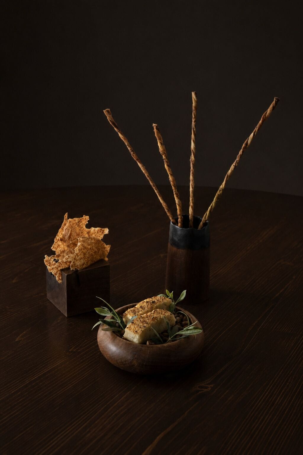 Una mesa de madera oscura con una caja cuadrada pequeña que contiene un alimento, un tazón de madera redondo con un alimento y hojas verdes, y un recipiente cilíndrico con palitos.