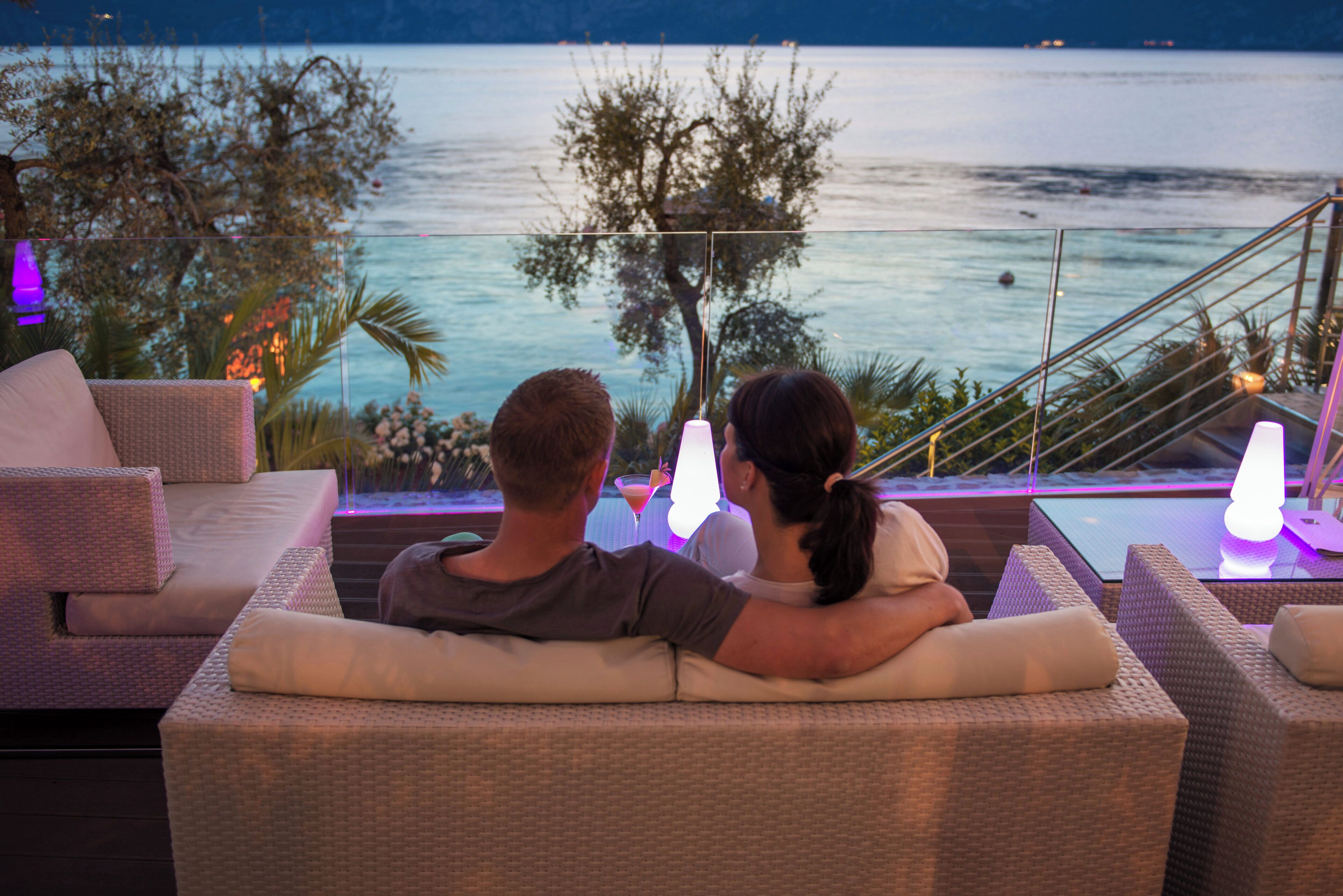 A couple sitting on a balcony overlooking a body of water, with outdoor furniture and ambient lighting.