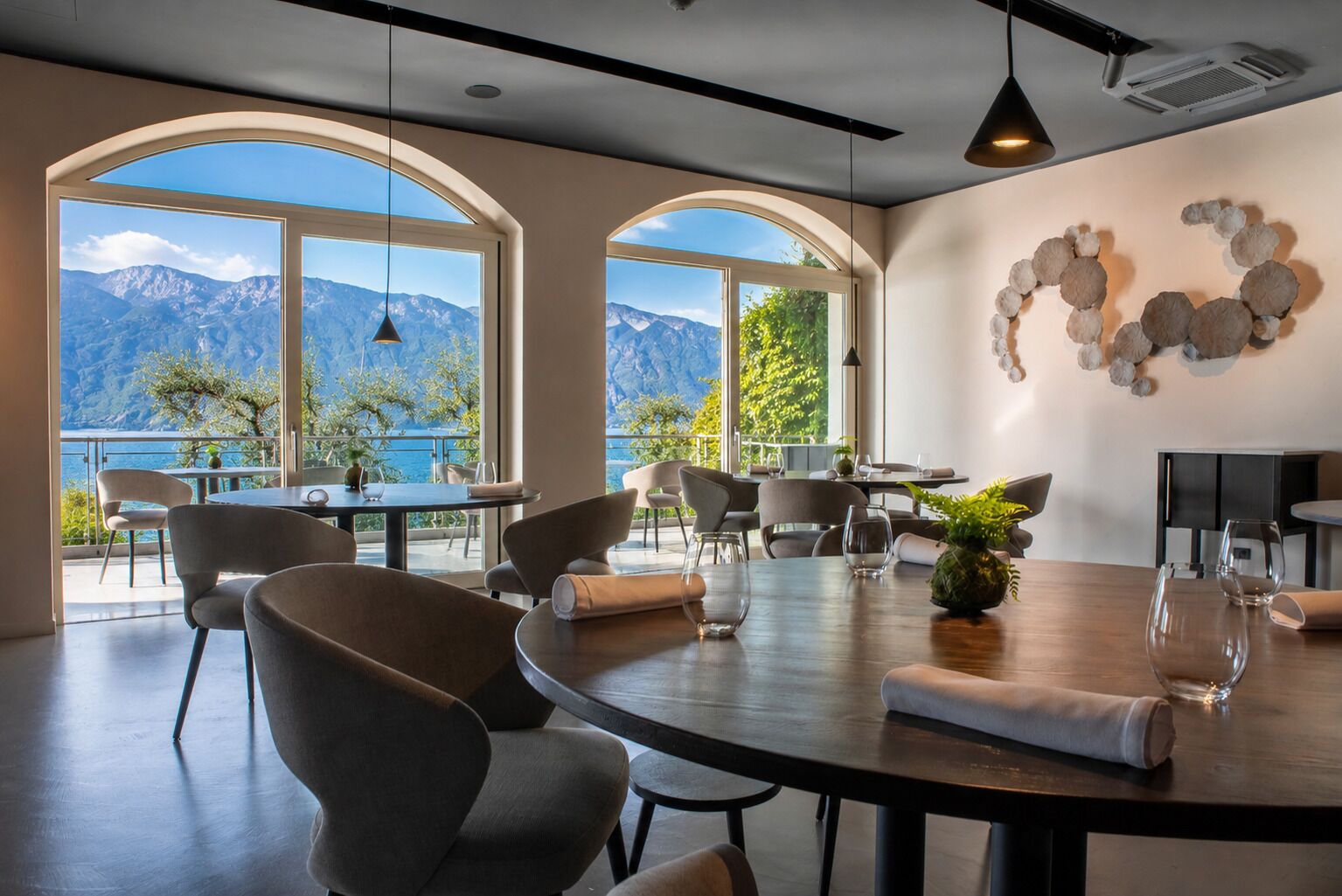 A dining area with a view of mountains and a body of water, featuring round tables, chairs, and a decorative wall piece.