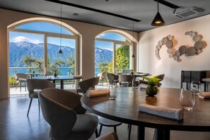 Un espace repas offrant une vue sur les montagnes et un plan d’eau, avec des tables rondes, des chaises et un élément décoratif mural.
