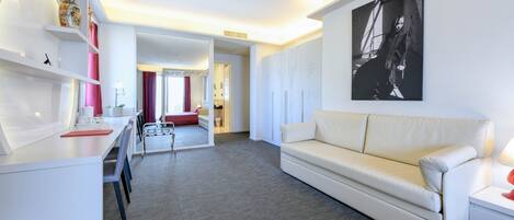 Ein modernes Hotelzimmer mit einem weißen Sofa, einem Schreibtisch mit Stuhl, einem kleinen Tisch mit einem roten Buch und einem großen gerahmten Bild an der Wand.