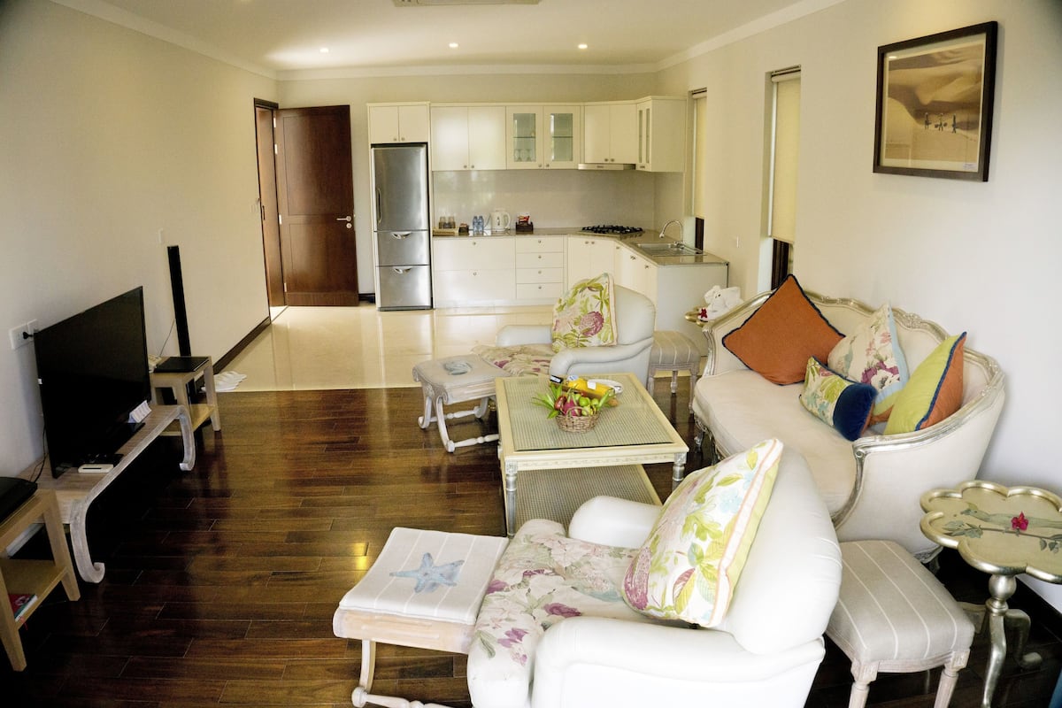 A modern living room with a flat-screen TV, a sofa with floral cushions, a dining table with a fruit basket, and a kitchen in the background.