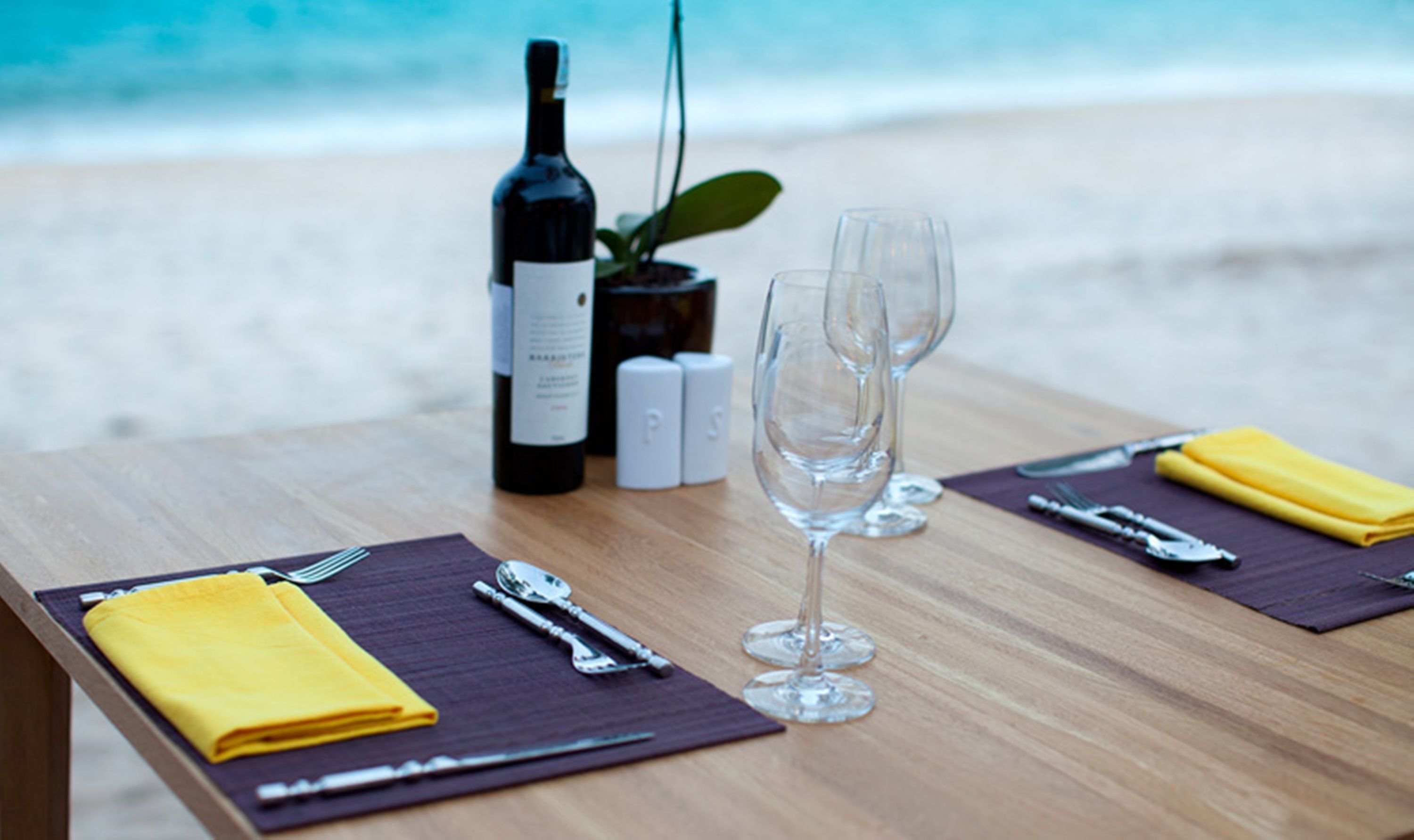 A table set with a bottle of wine, two glasses, cutlery, and a yellow napkin on a wooden table by the beach.