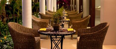 A row of wicker chairs and tables in a covered outdoor area with plants and columns.