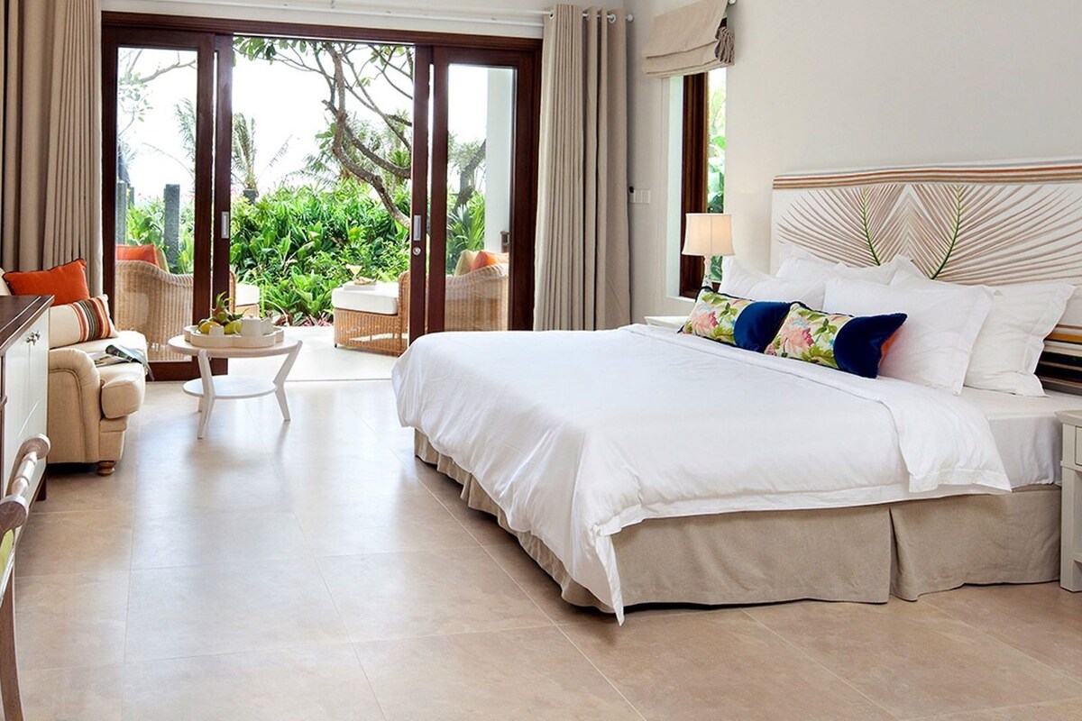 Habitación de hotel con una cama grande, una mesita de noche con lámpara, una silla, una pequeña mesa con una planta y vistas al exterior con vegetación.