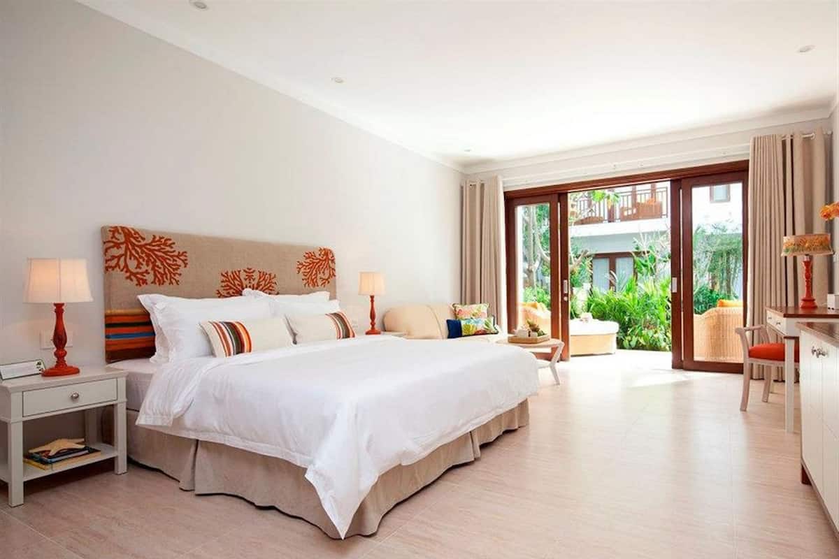 A spacious bedroom with a large bed, two bedside tables, and a view of a lush garden through a sliding glass door.