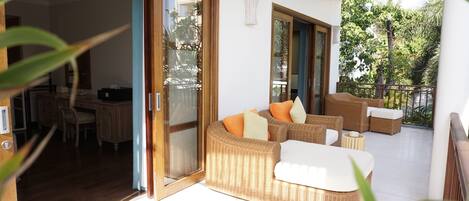 Un salón moderno con muebles de mimbre, un ventanal de vidrio que da a un balcón y un comedor visible a través del ventanal.