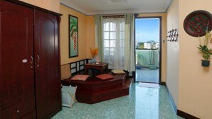 A room with a wooden wardrobe, a small table with a vase, a bench with cushions, and a view of the outside through a window. - La Suisse Hotel (Nha Trang)